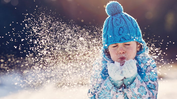 Pieni lapsi puhaltaa lunta käsistään sininen tupsupipo päässä.