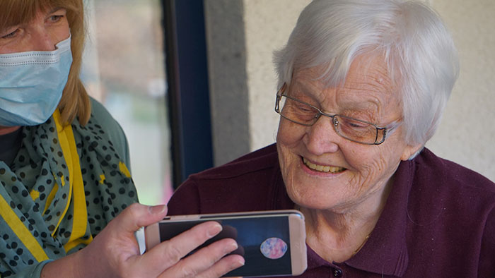Vanha nainen katsoo puhelinta hymyillen. Puhelinta pitelee keski-ikäinen nainen maski kasvoillaan.