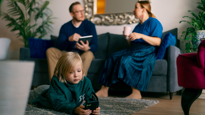 Sosiaalityöntekijä kotikäynnillä, istuu sohvalla äidin kanssa. Lapsi makaa lattialla ja katsoo puhelinta.